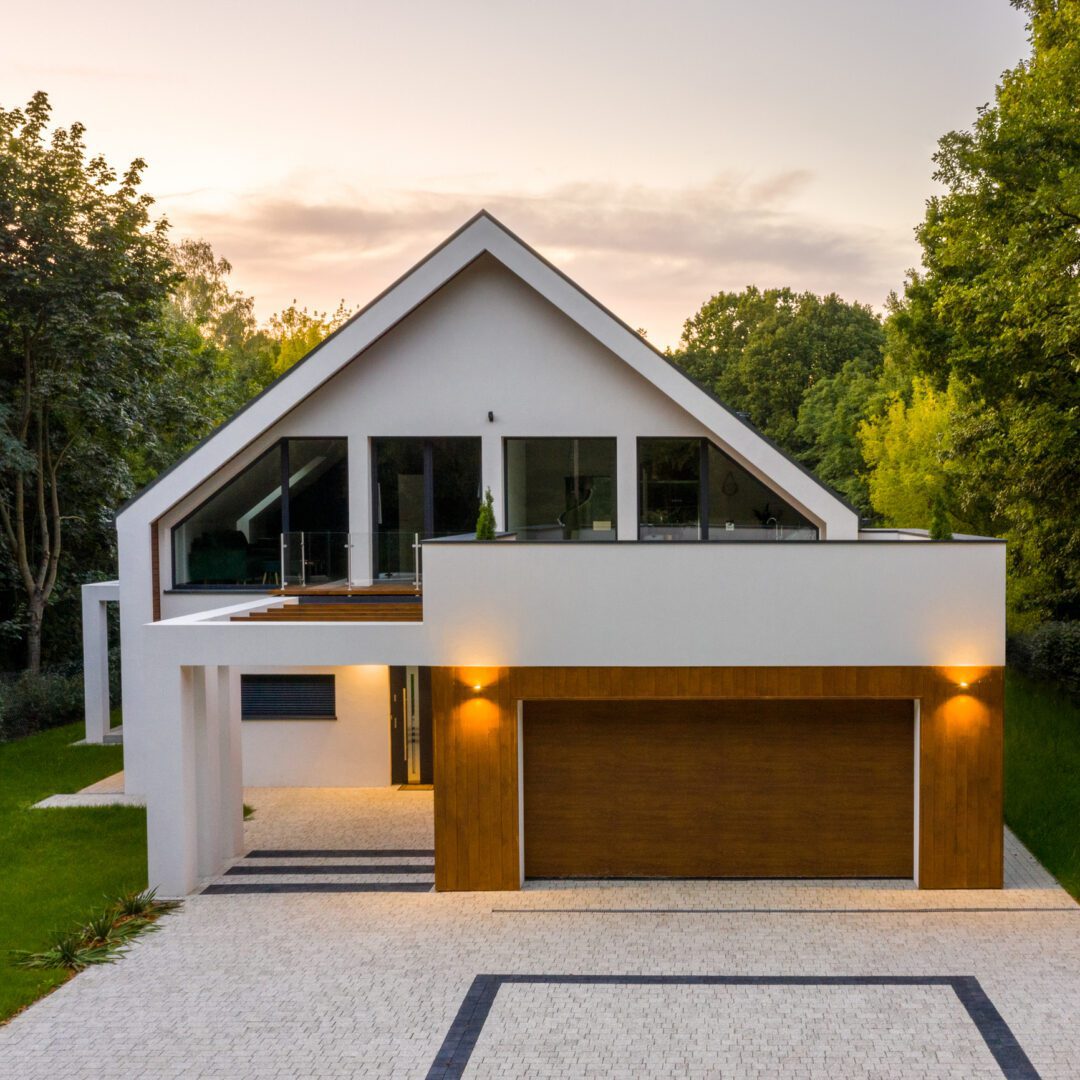 Sunset over elegant white house with forest around, big garage and cobblestone driveway, drone view