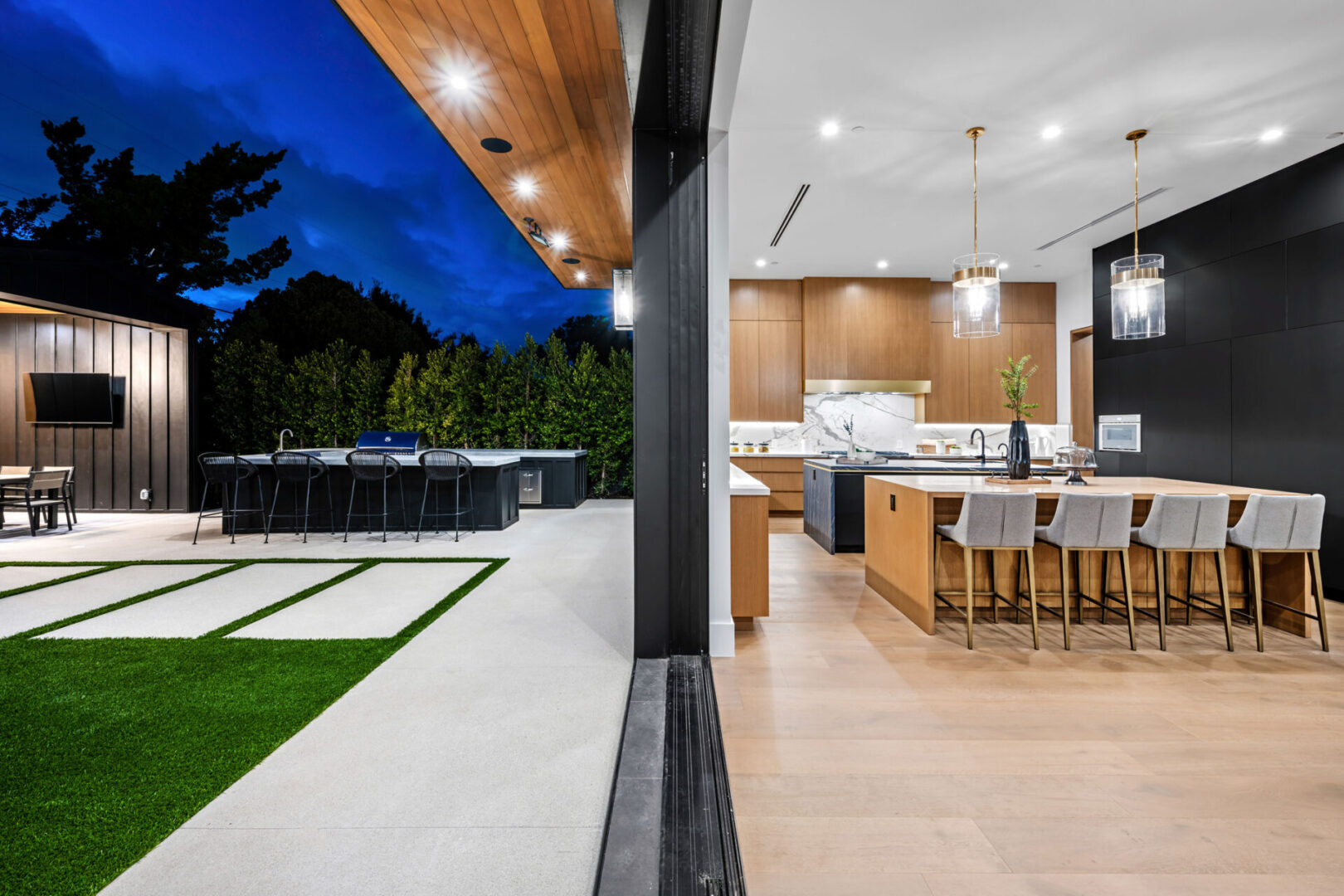 Modern indoor and outdoor kitchen with wooden accents, pendant lights, and a spacious patio with a grill and seating area at dusk.